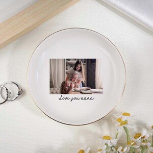 May include: White ceramic trinket dish with a gold rim, featuring a printed photo of a woman and a girl. The dish has the handwritten text "Love you nana". Two silver rings and daisies are also visible.
