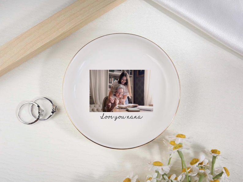 May include: White ceramic trinket dish with a gold rim, featuring a printed photo of a woman and a girl. The dish has the handwritten text "Love you nana". Two silver rings and daisies are also visible.