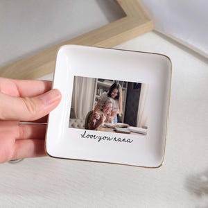 May include: A white square ceramic trinket dish with a gold rim. The dish features a photograph of a grandmother and granddaughter, with the handwritten text "Love you nana" below the photo.