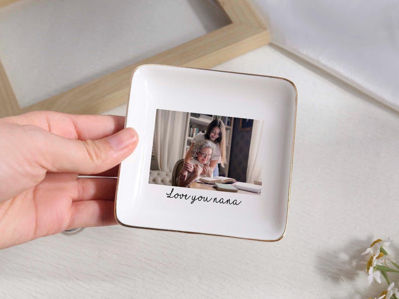 May include: A white square ceramic trinket dish with a gold rim. The dish features a photograph of a grandmother and granddaughter, with the handwritten text "Love you nana" below the photo.