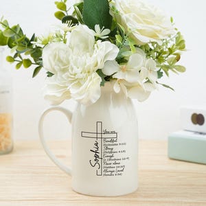 May include: A white ceramic vase with a handle, filled with white flowers and greenery. The vase features a cross design with the name "Sophia" and inspirational text. A glass bottle and books are in the background.