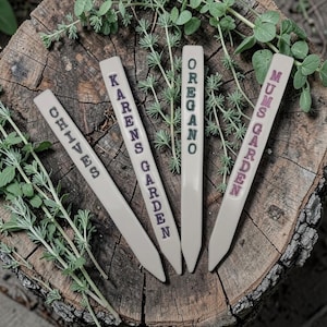 May include: Four cream-coloured garden markers with text in dark purple and green. The markers read "Chives", "Karens Garden", "Oregano", and "Mums Garden". The markers are arranged on a wooden surface with green foliage.