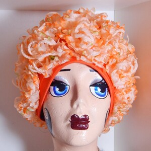 May include: A mannequin head with a bright orange fabric headband covered in orange and white fabric flowers. The mannequin has painted-on blue eyes, black eyeliner, and a dark red lipstick. The mannequin is wearing a silver rhinestone choker and a black feather boa.