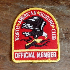 May include: Red, yellow and white embroidered patch with the text "North American Hunting Club" and a black and white eagle with a gun. The text "Official Member" is at the bottom of the patch.