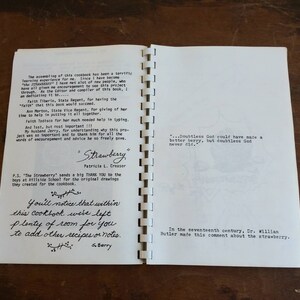 May include: Open cookbook with spiral binding, featuring handwritten text and printed content. The title "Strawberry" is visible, along with author's name. The pages include quotes and acknowledgements. The cookbook is open on a wooden surface.