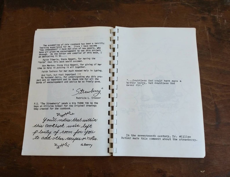 May include: Open cookbook with spiral binding, featuring handwritten text and printed content. The title "Strawberry" is visible, along with author's name. The pages include quotes and acknowledgements. The cookbook is open on a wooden surface.