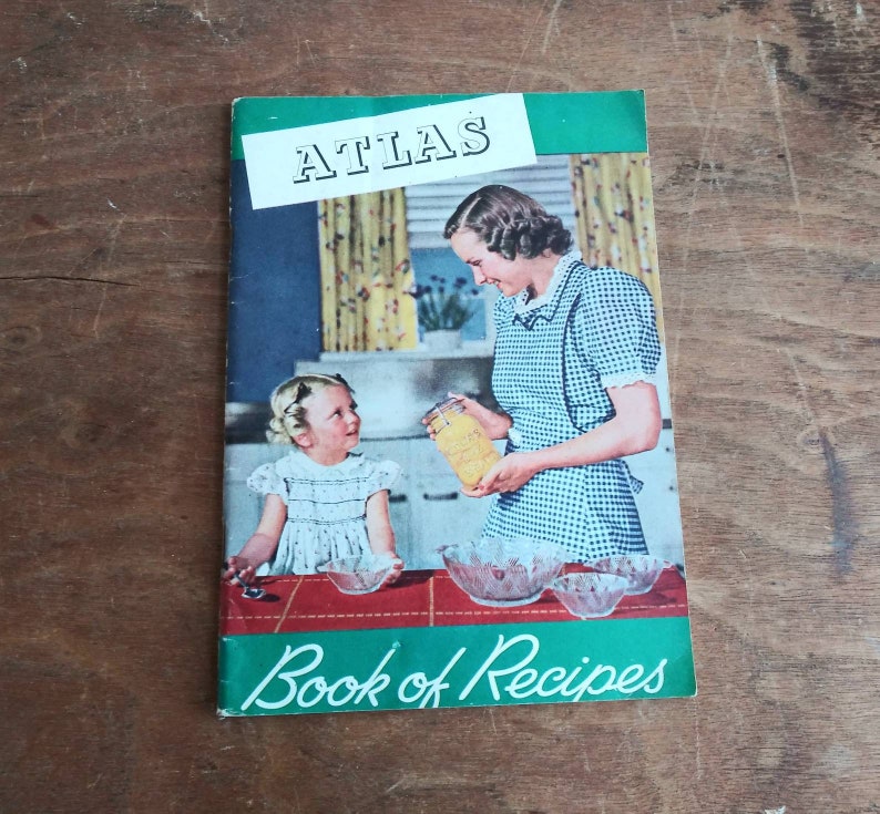 May include: A vintage green and white cookbook with the title "Atlas Book of Recipes". The cover features a woman and a young girl in a kitchen setting, with a jar of preserves and bowls on the counter.