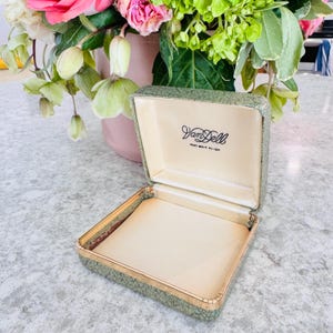 May include: Open vintage jewelry box with a textured green exterior and gold trim. The interior is cream-colored with the brand name "Van Dell" printed in black. The box is rectangular and appears to be made of a durable material.