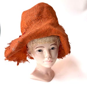 Può includere: Un cappello di iuta arancione con una tesa larga e bordi sfilacciati. Il cappello è posto su una testa di manichino con capelli biondi e lineamenti dipinti. Il cappello ha un aspetto rustico e fatto a mano.