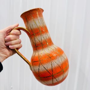 May include: A vintage orange and white ceramic pitcher with a gold handle and accents. The pitcher has a bulbous body and a tall, slender neck. The design features vertical white stripes with orange horizontal stripes and gold lines.