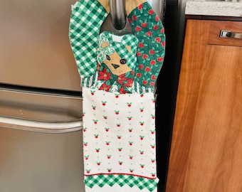 May include: A green and red patchwork kitchen towel with a bear applique hanging on a silver refrigerator handle. The towel has a white background with red polka dots and a green and white checkered border.