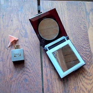 Puede incluir: Un estuche compacto rectangular abierto con un espejo y una sombra de ojos redonda, junto a un pequeño frasco de perfume con la letra "M". Un pequeño embudo de papel rosa está junto al frasco de perfume. Los artículos están sobre una superficie de madera.