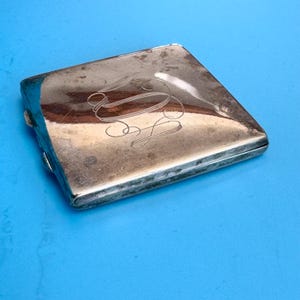 Könnte beinhalten: Ein rechteckiger, silberfarbener Kompaktspiegel mit Klappdeckel. Der Kompaktspiegel hat ein eingraviertes Monogramm auf der Oberseite. Die Kanten weisen Gebrauchsspuren auf, was darauf hindeutet, dass es sich um einen antiken oder Vintage-Artikel handelt.