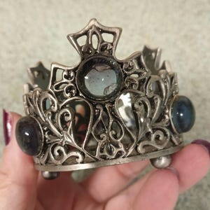 May include: A silver-toned metal crown-shaped holder featuring intricate scrollwork and filigree patterns. The design includes a central cross-shaped element and is accented with three smooth, dark, rounded cabochon stones set into the metal frame.