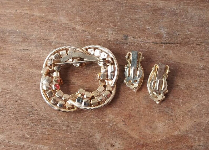 May include: A gold-tone brooch and matching clip-on earrings set with clear and red rhinestones. The brooch is shaped like a knot and the earrings are oval.
