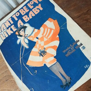 May include: A vintage sheet music cover with a blue background and a woman in a striped orange and white dress. The title of the song is "I Want to Be Loved Like a Baby" and the words and music are by William Witol.