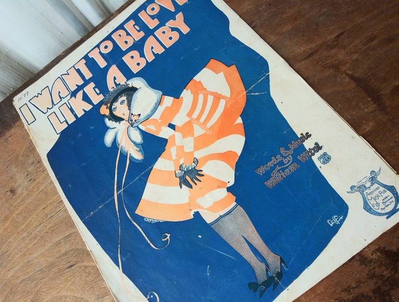 May include: A vintage sheet music cover with a blue background and a woman in a striped orange and white dress. The title of the song is "I Want to Be Loved Like a Baby" and the words and music are by William Witol.