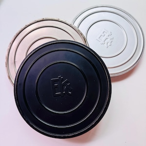 May include: Three circular film canisters in silver, black, and white. Each canister lid has a raised, embossed design in the center. The black canister is in the foreground, with the silver and white canisters behind it.
