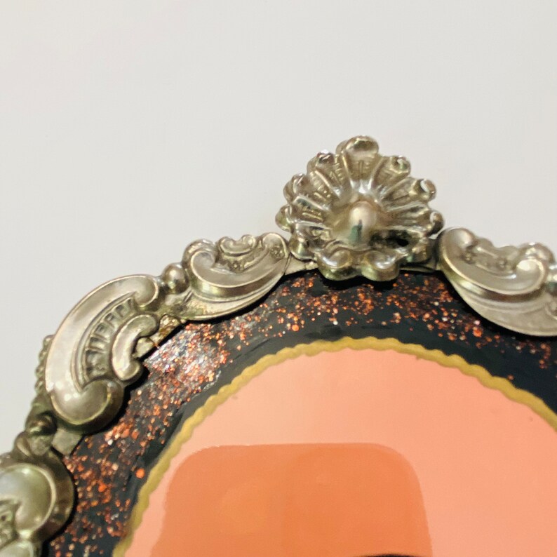 May include: A silver-toned ornate frame with a black and orange glitter background and a pink interior. The frame has a decorative design with a scalloped top.