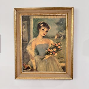 May include: A framed painting of a woman in a blue dress holding a bouquet of pink and white roses. The woman is standing in front of a window with a view of a garden. The painting is signed "C. Gray" in the bottom right corner.