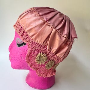 May include: A vintage-style pink satin bonnet featuring a pleated crown and a wide, intricate crocheted lace border. The lace trim is accented with two beige, circular floral-shaped appliqués on the side, providing a textured contrast.