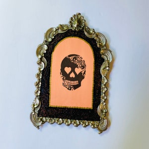 May include: A black and white sugar skull with hearts for eyes is printed on a peach-colored card inside a silver ornate frame.