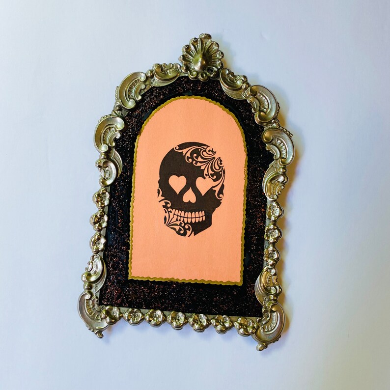 May include: A black and white sugar skull with hearts for eyes is printed on a peach-colored card inside a silver ornate frame.