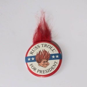 May include: A red, white, and blue button with a cartoon troll with red hair and a smiling face. The button says "Russ Troll for President" with three stars on each side of the text.