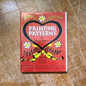 以下が含まれることがあります： 表紙にハート型のデザインが施された赤い本。本のタイトルは「Painting Patterns for Home Decorators」で、著者名はRuth Wyeth Spears。本の表紙には、花柄のデザインと黄色のリボンも描かれている。