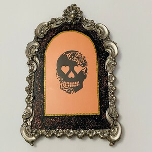 May include: A silver ornate frame with a black and pink glitter background. A black and white silhouette of a skull with hearts for eyes is in the center of the frame.