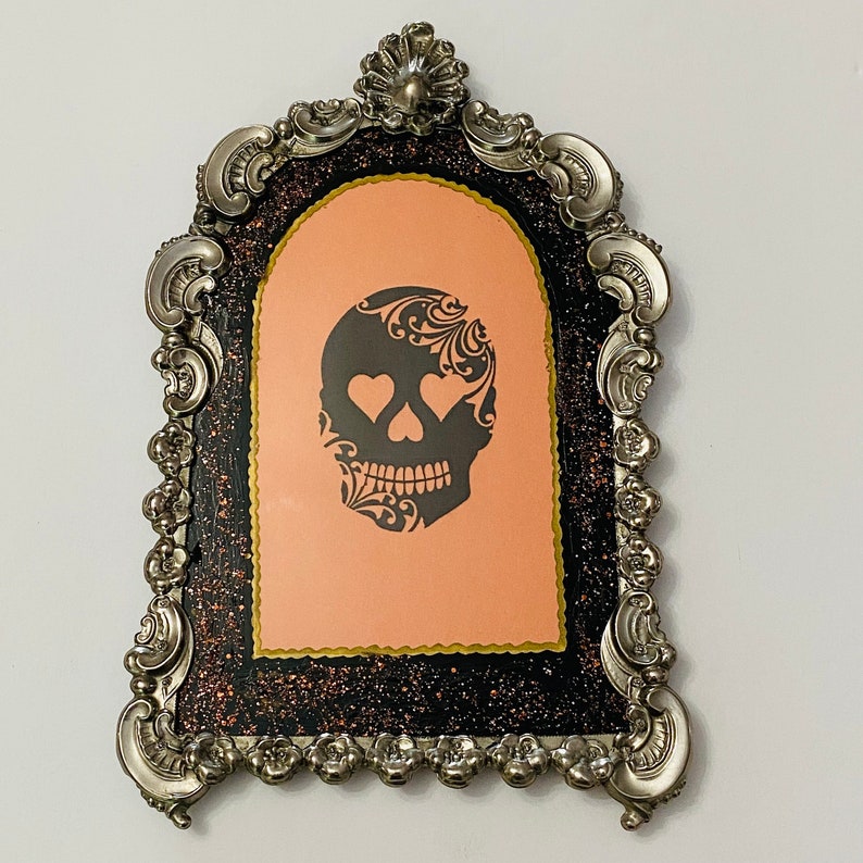May include: A silver ornate frame with a black and pink glitter background. A black and white silhouette of a skull with hearts for eyes is in the center of the frame.