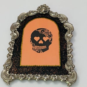 May include: A silver ornate frame with a black and orange glitter background. The frame holds a peach-colored card with a black and white sugar skull design with hearts for eyes.