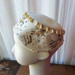 May include: A white felt beret with a beaded trim. The trim features pearls, sequins, and small beads in a variety of shapes and sizes.
