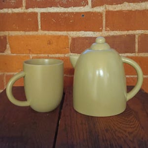 Könnte beinhalten: Ein hellgrünes Keramik-Teeservice mit einer passenden Tasse. Die Teekanne hat einen abgerundeten Körper, einen gebogenen Griff und einen kleinen Ausguss. Die Tasse hat ein einfaches, abgerundetes Design mit einem gebogenen Griff.