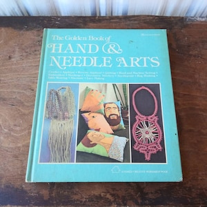 Könnte beinhalten: Ein Vintage-Buch mit dem Titel "The Golden Book of Hand & Needle Arts" mit türkisfarbenem Einband. Das Cover zeigt Bilder verschiedener Handarbeiten, darunter Häkeln, Sticken und Makramee. Das Buch ist ein Familien-Kreativ-Workshop-Buch.
