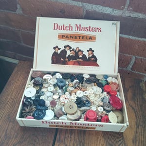 May include: A vintage box of assorted buttons, featuring a variety of colors, sizes, and materials. The box is labeled "Dutch Masters PANETELA" with an image of people in hats. The box is open, revealing the buttons inside.