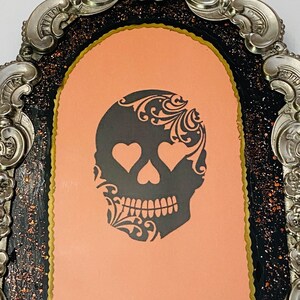 May include: A black and white skull with a floral design and heart-shaped eyes is displayed in a silver ornate frame with a black and gold background.