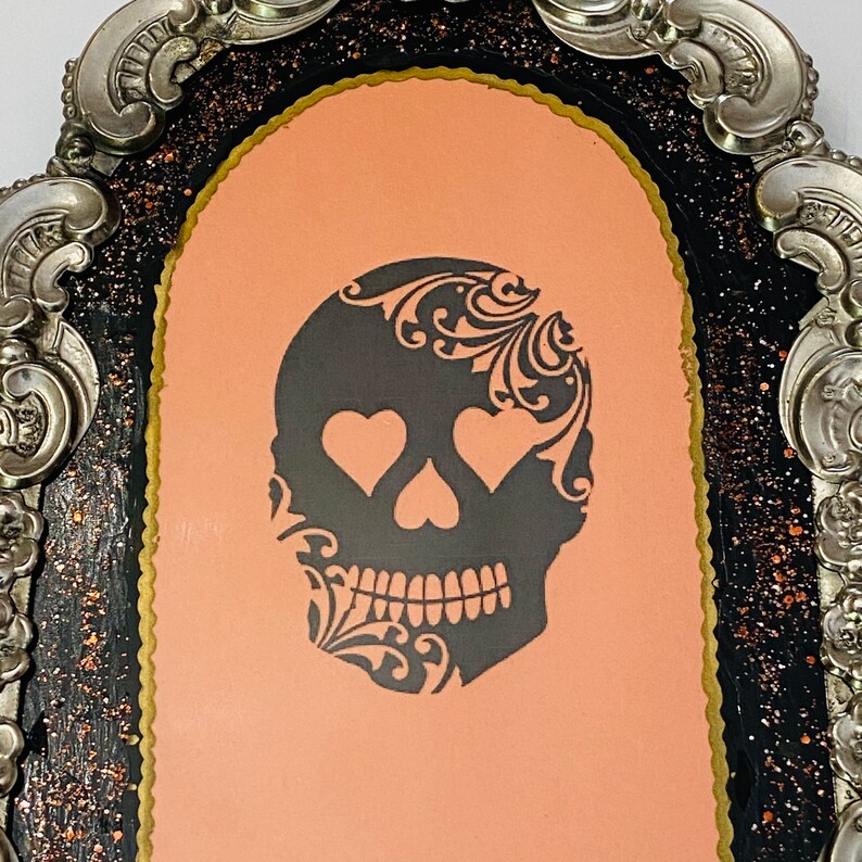May include: A black and white skull with a floral design and heart-shaped eyes is displayed in a silver ornate frame with a black and gold background.