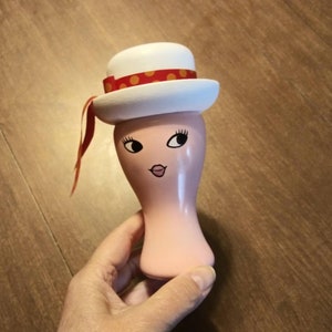 May include: A pink ceramic figurine with a white hat with red polka dots and a red ribbon. The figurine has a painted face with black eyes, a pink nose, and a red mouth.
