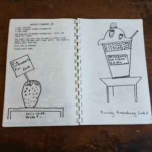 May include: A spiral-bound notebook open to two pages of hand-drawn illustrations and text. One page features a strawberry with a sign reading "Strawberry For Sale." The other page depicts a strawberry ice cream soda. Text includes a recipe for uncooked strawberry jam.
