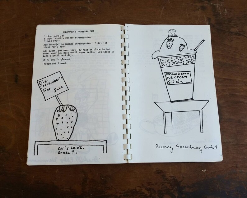 May include: A spiral-bound notebook open to two pages of hand-drawn illustrations and text. One page features a strawberry with a sign reading "Strawberry For Sale." The other page depicts a strawberry ice cream soda. Text includes a recipe for uncooked strawberry jam.