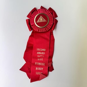 May include: A red satin award ribbon featuring a pleated circular rosette at the top with a gold-toned center emblem displaying a tree icon and the text Telephone Pioneers of America. The long tail is printed with Second Award Life Pioneer Hobby Show.