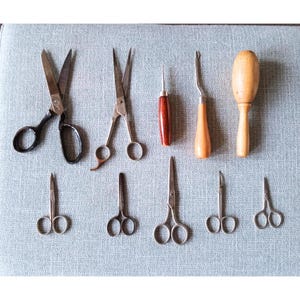 May include: A collection of nine sewing tools, including scissors, awls, and a thimble. The tools are arranged on a light blue fabric background.