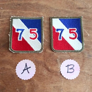 Può includere: Due toppe militari vintage con un designo bianco, rosso e blu. Le toppe sono rettangolari e hanno il numero "75" ricamato in rosso su sfondo bianco. Le toppe sono etichettate "A" e "B".