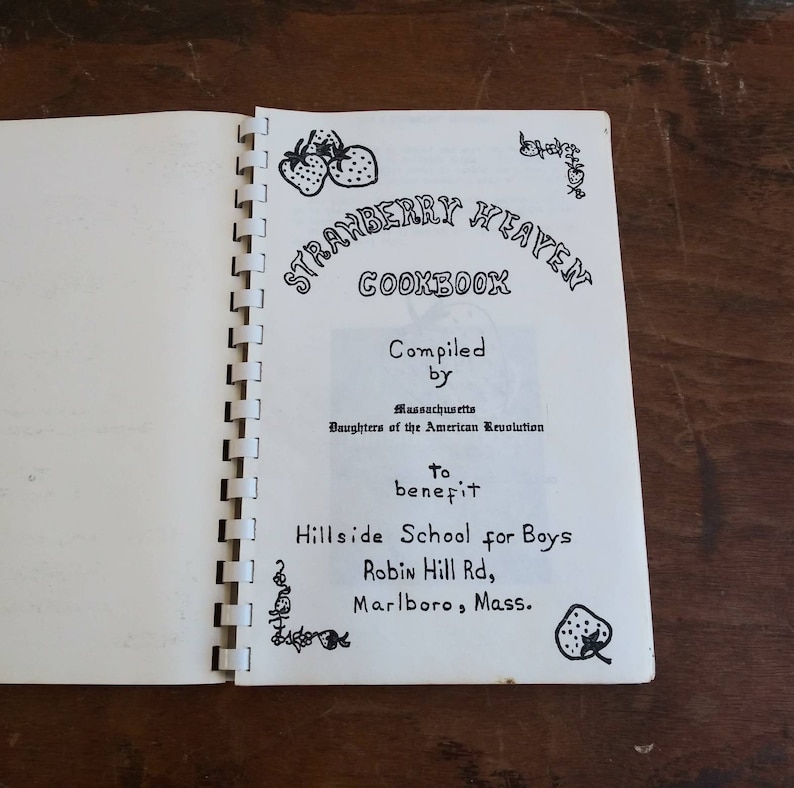 May include: Open cookbook titled "Strawberry Heaven Cookbook" with spiral binding. The cover features handwritten text and illustrations of strawberries. The book was compiled by the Massachusetts Daughters of the American Revolution to benefit Hillside School for Boys.