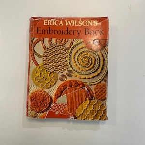 May include: A vintage book titled "ERICA WILSON'S Embroidery Book" with a colourful cover featuring various embroidery stitches and patterns in shades of orange, yellow, and brown.