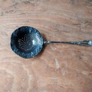 Puede incluir: Un colador de té de plata con un cuenco redondo perforado y un mango largo. El cuenco está decorado con un diseño de vid.