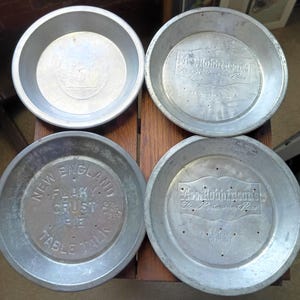 Może przedstawiać: Cztery zabytkowe aluminiowe formy do ciasta. Dwie formy są oznaczone jako "Mrs. Robinson's Fine Restaurant Pies", jedna jako "New England Flaky Crust Pie Table Talk", a jedna jako "Bakers Square".