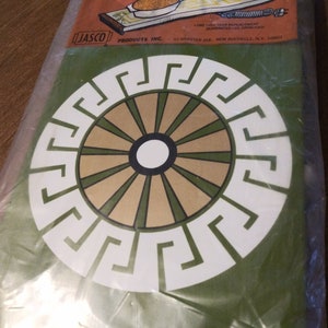 Puede incluir: Un posavasos circular verde y blanco con un borde de clave griega y un diseño de sol naciente en el centro. El posavasos está empaquetado en plástico.