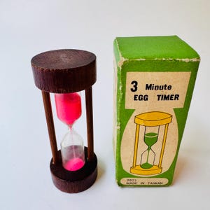 May include: A wooden hourglass with bright pink sand and a matching green cardboard box. The box features the text 3 Minute EGG TIMER and MADE IN TAIWAN. The hourglass has dark brown circular wooden ends connected by thin vertical wooden supports.